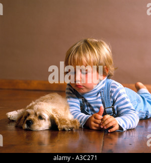 Little Boy indossando tute sdraiato sul pavimento di legno con il suo cucciolo Cocker Spaniel accanto a lui Foto Stock