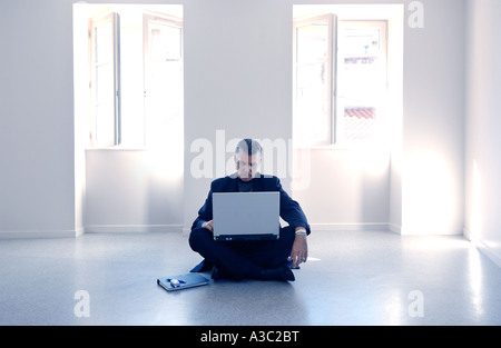 Uomo con notebook computer seduto sul pavimento da solo nel mezzo di un bianco stanza vuota Foto Stock