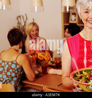 Ritratto di una donna matura in possesso di una ciotola di insalata e i suoi amici seduti al tavolo da pranzo in background Foto Stock