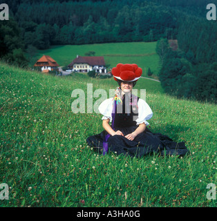 Foresta Nera Ragazza con cappello tradizionale seduta in un prato con una fattoria in background Foto Stock