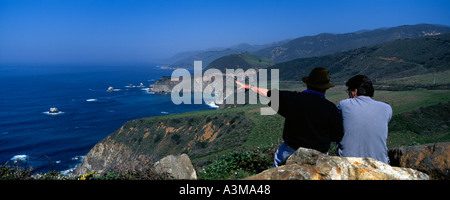 Due uomini seduti sulla roccia che si affacciano sull'Oceano Pacifico costa vista sud di Big Sur in California con Bixby Bridge e Big Creek Foto Stock