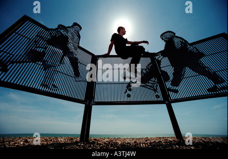 Silhouette di un giovane uomo appollaiato sulla cima di Modern scherma sulla spiaggia di ciottoli con mare in background. Foto Stock