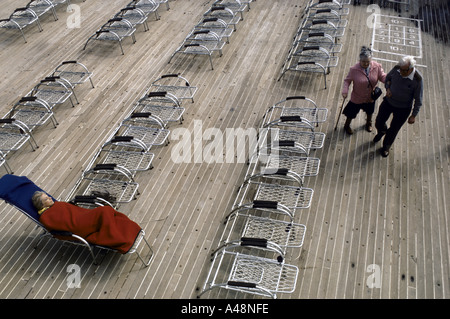 Coppia di anziani camminando sul ponte come donna sdraiata sulla piegatura letto sun mantiene caldo sotto una coperta durante qe2 vacanza in crociera Foto Stock