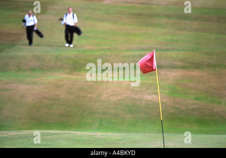 Un rosso golf bandiera in una messa a fuoco nitida soffia il vento come due giovani golfisti a piedi lungo il fairway per la loro ripresa successiva Foto Stock