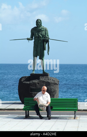 Uomo anziano giornale di lettura sul banco di essere guardato da un ANATERVE Guanche statua in Candelaria Tenerife Canarie Spagna Foto Stock