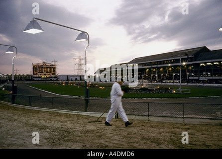 Un assistente maschio indossa una caldaia bianco tuta rastrellando la via per il Greyhound Racing come il sole tramonta su Walthamstow Stadium Foto Stock