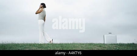 Donna che cammina su erba parlando al cellulare, valigetta metallica sul terreno dietro di lei Foto Stock