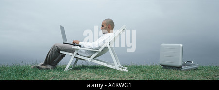 Uomo seduto in poltrona con il computer portatile in giro, vista laterale, aperto valigetta metallica sul terreno dietro di lui Foto Stock