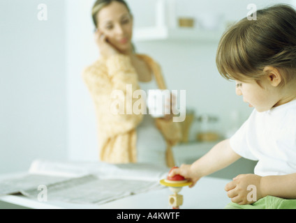 Bambina giocando con il giocattolo, donna holding mug e parlare al telefono in background Foto Stock