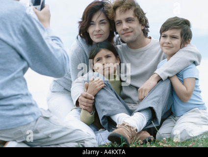 I genitori con un ragazzo e una ragazza seduta su erba che posano per una foto, ragazzo prendendo foto Foto Stock