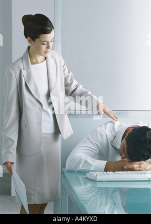 Uomo dorme da scrivania, collega di sesso femminile in piedi accanto a lui toccando la sua spalla Foto Stock