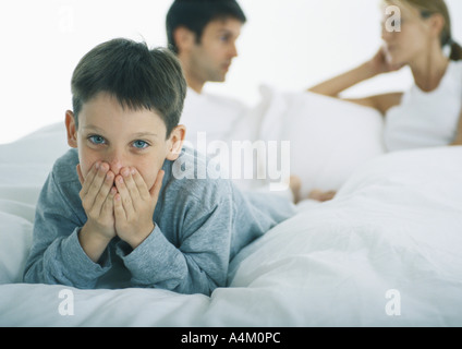 Ragazzo disteso sul ventre nel letto che copre la bocca con le mani, i genitori a discutere in background Foto Stock