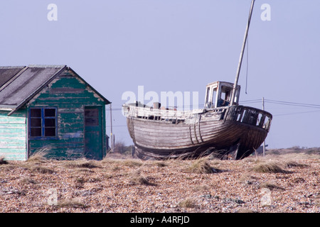 Abbandonata la pesca in legno barca accanto ad un verde fatiscente capanna sulla spiaggia di ciottoli a Dungeness nel Kent. Foto Stock