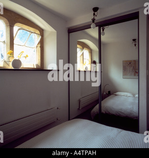 Un grande specchio sulla cabina armadio nella piccola camera da letto bianca con finestra alta Foto Stock