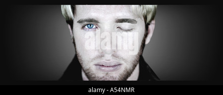 Capelli biondi uomo con barba lunga faccia si chiude un occhio e apre il suo altro blue eye in un surreale pongono contro uno sfondo scuro Foto Stock