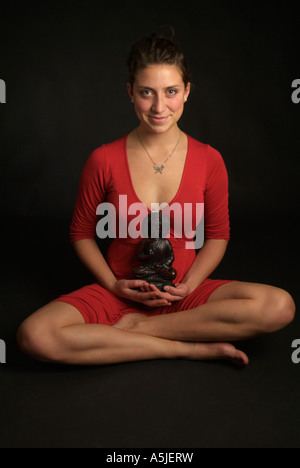 Studio ritratto di una giovane donna con sapendo sorriso in un abito rosso seduta zampe trasversale tenendo un buddha figura per il suo torace Foto Stock