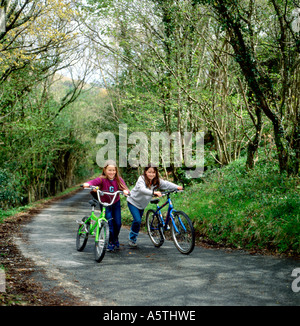 Felici ragazze pre-adolescenti giovani ciclisti bambini che spingono le biciclette su una collina su una corsia di campagna in primavera a Carmarthenshire Galles Regno Unito KATHY DEWITT Foto Stock