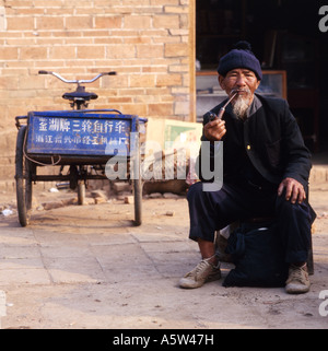 Anziani locali barbuto uomo si siede accanto al suo triciclo fumare la pipa,nella provincia dello Yunnan il sud-ovest della Cina. Foto Stock