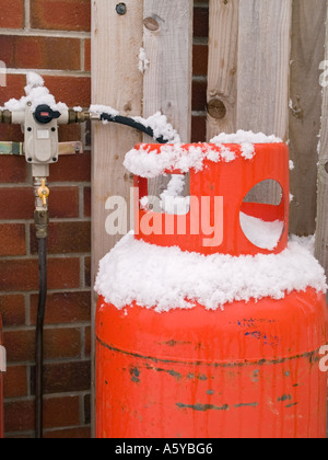 Domestico calor rosso Bottiglia di gas collegato alla valvola su "muro di mattoni' con neve invernale sul cilindro, recinzione di legno dietro. Foto Stock