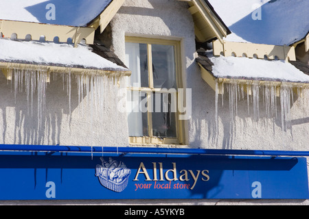 Tetto coperto di neve spiovente coperto di iciclette a punta dura nel negozio Alldays in Braemar Scozia UK. Il sole di febbraio con i icicles appesi ai tetti Foto Stock