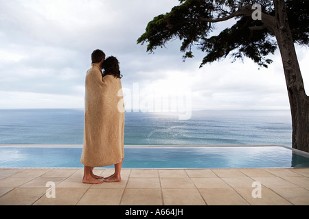 Giovane coperto in un asciugamano in piedi dalla piscina infinity, vista laterale Foto Stock