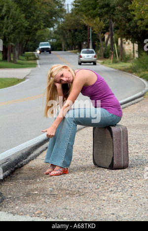 Giovane ragazza bionda con i capelli lunghi che indossa jeans seduto su una valigia di fianco alla strada cercando un modo fuori città Foto Stock