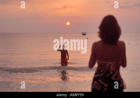 Il colore di un'immagine orizzontale di paunchy un uomo di mezza età in costume da bagno nell'oceano al tramonto sventolano fuori per una donna sulla riva Foto Stock