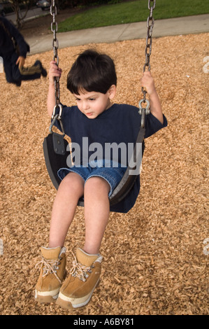 Colore immagine verticale di un bambino che indossa una maglietta blu jean corti e camoscio scarpe da trekking basculante in un parco giochi swing Foto Stock