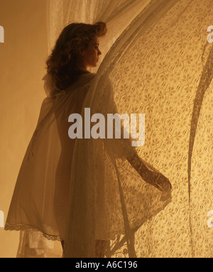 Donna in nightgown guardando fuori camera da letto finestra ventoso Foto Stock