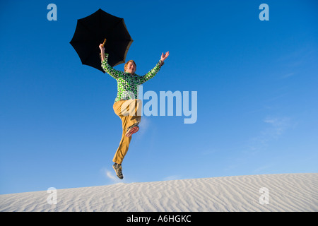 young man holding umbrella while leaping over sand dune Foto Stock