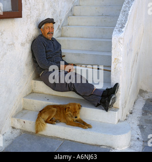 Anziani carattere locale in berretto con bastone da passeggio si siede sulla pietra dipinta di fasi con il suo cane in Oia Santorini Isole greche Foto Stock