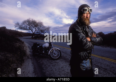 Uomo in giacca di pelle e cappuccio in piedi nella parte anteriore della sua moto su strada a vuoto nei pressi di albero in California modello rilasciato image Foto Stock