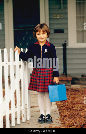 Bambina di cinque anni vestite per asilo in piedi accanto a un bianco Picket Fence e tenendo la sua scatola di pranzo Foto Stock