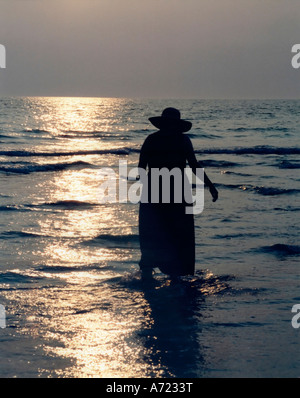 Silhouette di donna in abito e cappello che guadi attraverso l'acqua poco profonda su una spiaggia di oceano in Florida al crepuscolo Foto Stock