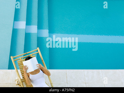 Donna seduta in sedia a sdraio in piscina, lettura, ad alto angolo di visione Foto Stock