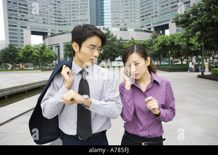 Due giovani dirigenti aziendali in office park, donna tramite telefono cellulare e uomo tenendo su braccio con orologio Foto Stock