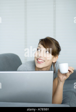 Giovane donna sul lettino, utilizzando laptop e azienda tazza da caffè, guardando verso l'alto Foto Stock