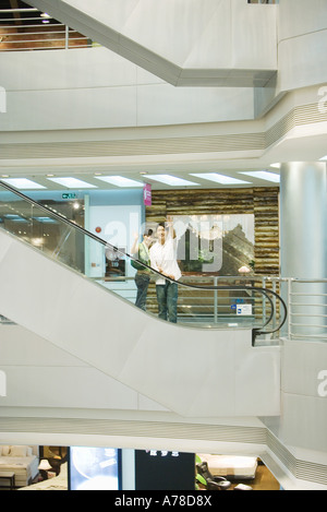 Giovane prendendo in escalator shopping mall, sventolando in telecamera, a metà distanza Foto Stock