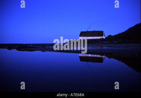 Tetto di Paglia Tradizionali Cottage irlandese con luci accese dentro la casa con la sua riflessione in acqua sotto un cielo di sera Foto Stock