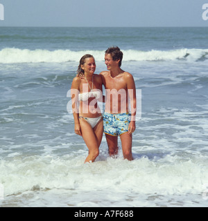 Coppia giovane camminando a braccetto nel surf lungo la riva del mare sulla spiaggia di sabbia di Fajara in Gambia Africa occidentale Foto Stock