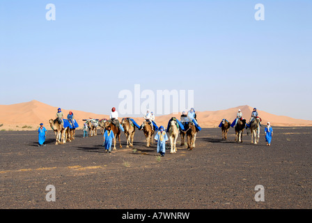 Gruppo di turisti passeggiate sui cammelli attraverso il piatto deserto roccioso reg serir con sanddunes in background Erg Chebbi Merzouga Moro Foto Stock