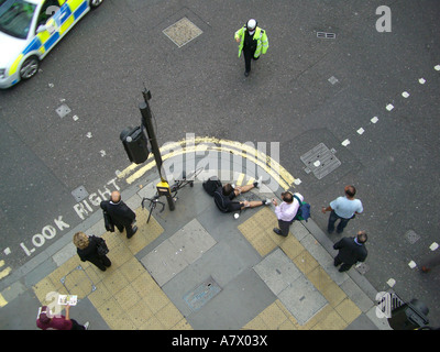 Vista aerea del ciclista steso sul marciapiede che riceve cure con l'agente di polizia e testimone dopo l'incidente sulla City of London Street Foto Stock