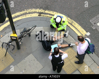 Vista aerea del ciclista steso sul marciapiede che riceve cure con l'agente di polizia e testimone dopo l'incidente sulla City of London Street Foto Stock