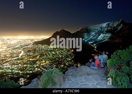 Un gruppo di escursionisti sedersi sul vertice della testa di leone ammirando la Table Mountain e la città di Città del Capo in una notte di luna. Foto Stock