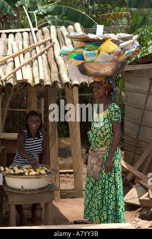 Donna del villaggio di cottura snack banana su fuoco di carbone con street hawker amico vendere beni dalla coppa portati sulla sua testa, Ghana Africa occidentale Foto Stock