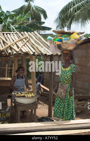 Donna del villaggio di cottura snack banana su fuoco di carbone con street amico commerciante vendita di merci dalla coppa portati sulla sua testa, Ghana Africa occidentale Foto Stock