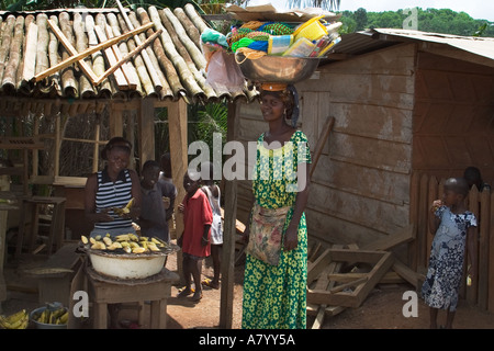 Donna del villaggio di cottura snack banana su fuoco di carbone con street amico commerciante vendita di merci dalla coppa portati sul suo capo, Ghana, Africa occidentale Foto Stock