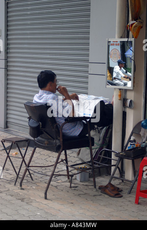 Barbiere leggendo il giornale sulla strada a Saigon Vietnam Foto Stock
