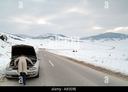 Giovane donna guardando sotto il cofano della sua auto dopo essere stati bloccati su una strada isolata in inverno dopo una pausa verso il basso Foto Stock