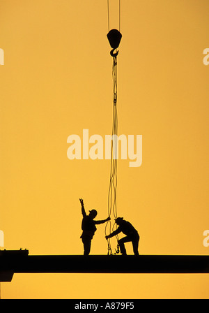 Silhouette di due lavoratori edili in piedi su una trave come una gru si abbassa alle prese ganci Foto Stock